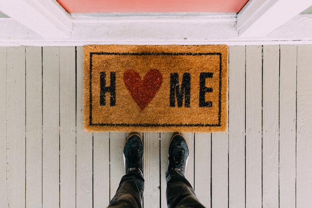 person standing in front of front door brown mat that says "home" with a red heart instead of an "o"